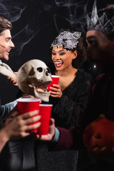 Cheerful multiethnic friends in halloween costumes clinking plastic cups near scary scull on black — Stock Photo