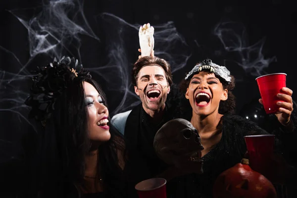 Riéndose africana americana mujer celebración juguete mano cerca excitado hombre durante halloween fiesta en negro - foto de stock