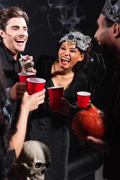Excited african american woman holding toy hand near multiethnic friends clinking plastic cups on halloween party on black — Stock Photo