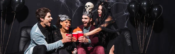 Amigos multiétnicos felices en trajes de Halloween tostadas con tazas de plástico en el sofá cerca de globos en negro, pancarta - foto de stock