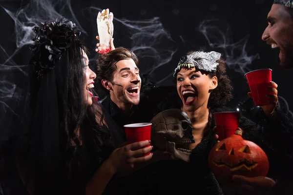 Laughing african american woman in halloween wolf mask scaring multiethnic friends with toy hand on black — Stock Photo