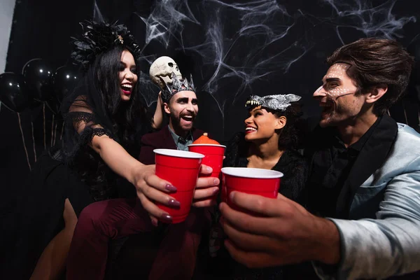 Happy multiethnic friends in halloween costumes toasting with plastic cups on black — Stock Photo
