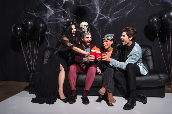 Multiethnische Freunde in Halloween-Kostümen, die mit Plastikbechern anstoßen, während sie auf dem Sofa neben Luftballons auf schwarz sitzen — Stockfoto