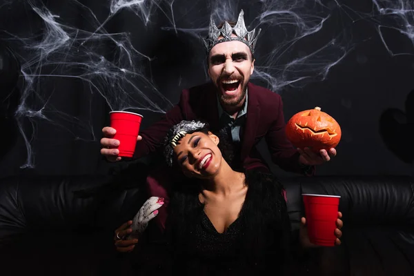 Riant homme en costume vampire halloween avec tasse en plastique et citrouille sculptée près de femme afro-américaine en masque de loup sur noir — Photo de stock