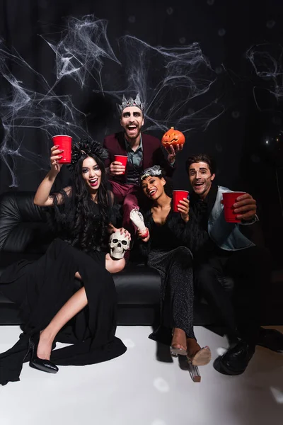 Amigos multiétnicos felices en disfraces de Halloween mirando a la cámara mientras se sienta en el sofá en negro - foto de stock