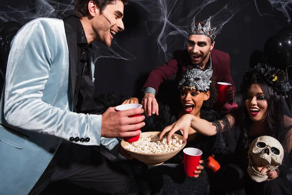 Thrilled interracial friends eating popcorn during halloween party on black — Stock Photo