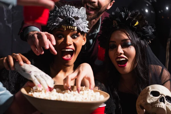 Excited interracial women in halloween costumes, with skull and toy hand near bowl with popcorn — Stock Photo