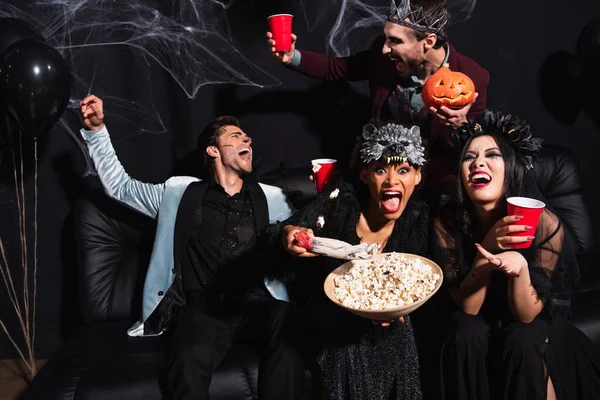 Thrilled multiethnic friends in halloween costumes eating popcorn and drinking beer during party on black — Stock Photo