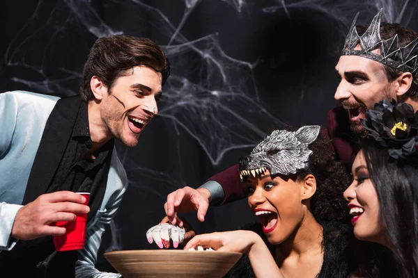 Asombrados amigos multiétnicos comiendo palomitas de maíz durante la fiesta de Halloween en negro - foto de stock