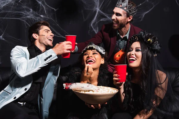 Hommes heureux griller avec tasse en plastique près des femmes interraciales en costumes d'Halloween manger du maïs soufflé sur noir — Photo de stock