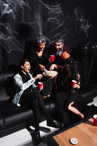 Високий кут зору багатоетнічних друзів у костюмах Хеллоуїна, які їдять попкорн біля декоративної павутинної мережі на чорному — стокове фото