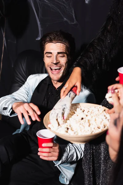 Man in halloween makeup laughing near woman taking popcorn with toy hand on black — Stock Photo