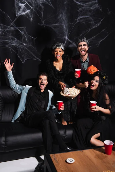 Mann winkt aufgeregten multiethnischen Freunden in Halloween-Kostümen zu, isst Popcorn und trinkt Bier auf Schwarz — Stockfoto