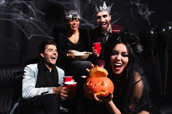 Animado ásia mulher mostrando esculpida abóbora perto rindo multicultural amigos no halloween trajes no preto — Fotografia de Stock