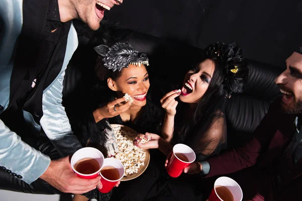 Excited interracial women in halloween costumes eating popcorn near men with beer on black — Stock Photo