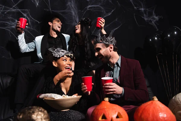 Amici multietnici in costumi di Halloween parlando mentre mangiano popcorn e bevono birra sul nero — Foto stock