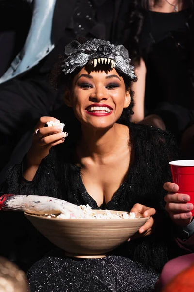 Excitada mujer afroamericana en máscara de lobo comiendo palomitas de maíz durante la fiesta de halloween en negro - foto de stock