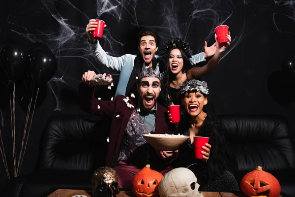 Thrilled multicultural friends in halloween costumes near popcorn, carved pumpkins and skulls on black — Stock Photo