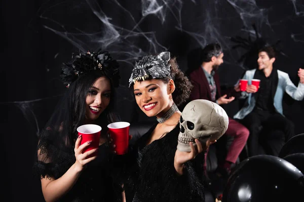 Cheerful interracial women with plastic cups and spooky skull near men talking on blurred and black background — Stock Photo