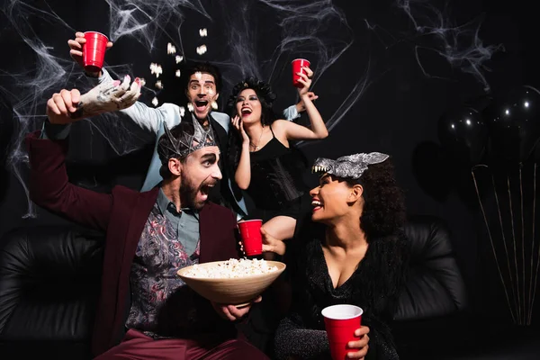 Excited man in vampire king crown scaring african american woman with toy hand near interracial friends on black — Stock Photo