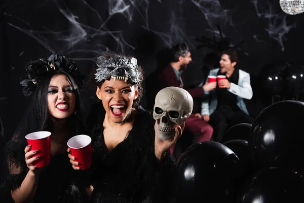 Міжрасові жінки в моторошних костюмах Хеллоуїна, що посміхаються і ростуть біля розмитих друзів, що говорять на чорному — стокове фото