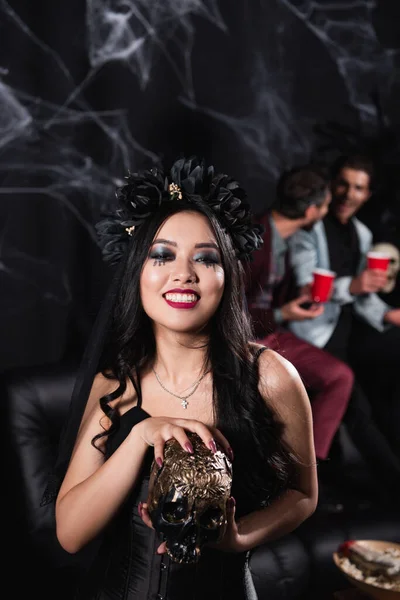 Smiling asian woman in vampire halloween costume holding spooky skull on black — Stock Photo