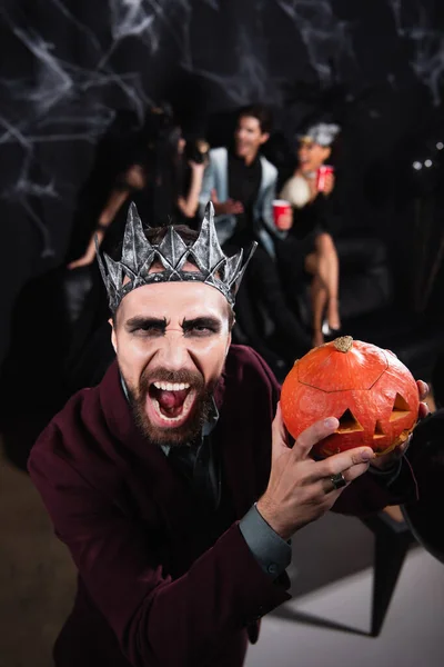 Enojado hombre en vampiro rey corona gruñendo en cámara mientras celebración halloween calabaza en negro - foto de stock