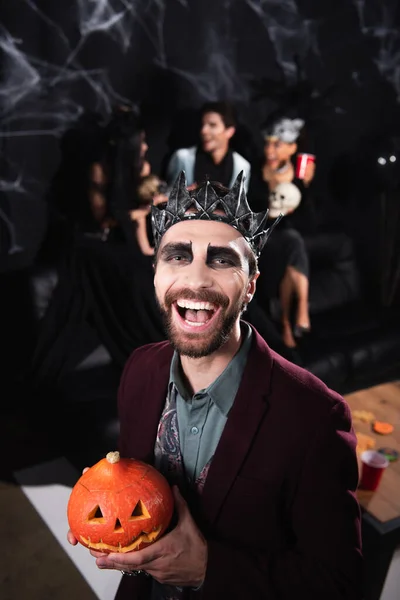 Cheerful man in vampire halloween costume laughing at camera while holding carved pumpkin on black — Stock Photo