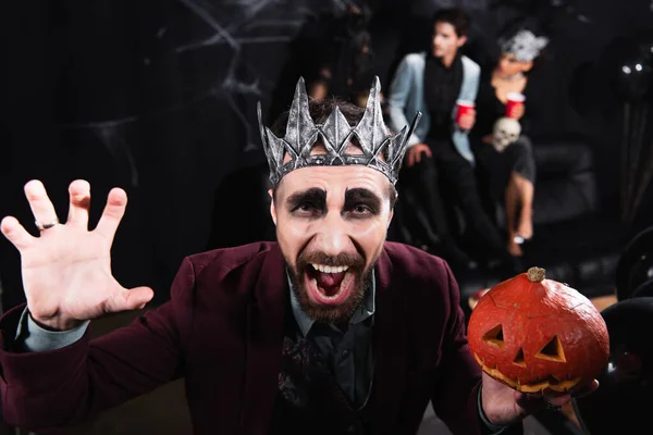 Hombre en vampiro rey corona celebración tallada calabaza y gruñendo en la cámara durante la fiesta de halloween en negro - foto de stock