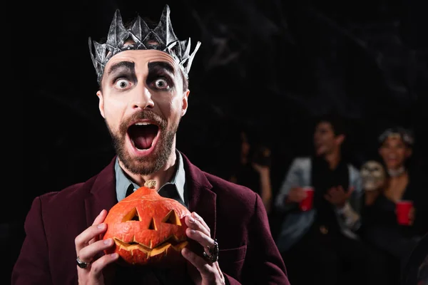 Aufgeregter Mann in Vampirkönigskrone mit gruseligem Halloween-Kürbis, während er auf schwarz in die Kamera schaut — Stockfoto