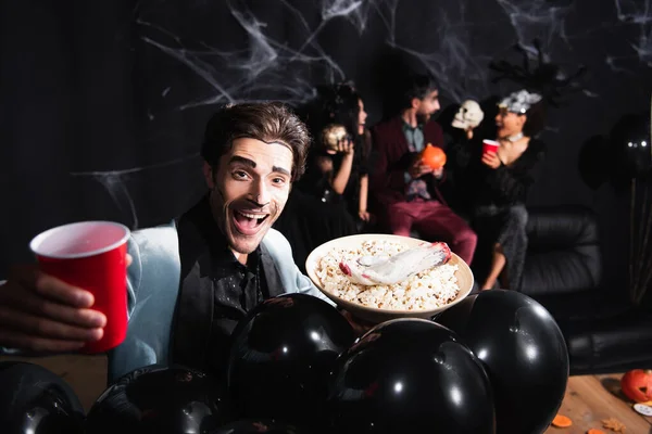 Feliz hombre en halloween maquillaje celebración de cerveza y palomitas de maíz cerca de globos negros y amigos interracial sobre fondo borroso y negro - foto de stock