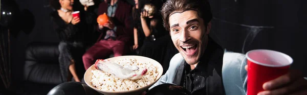 Cheerful man with halloween makeup holding bowl with popcorn and toy hand near blurred friends on black background, banner — Stock Photo
