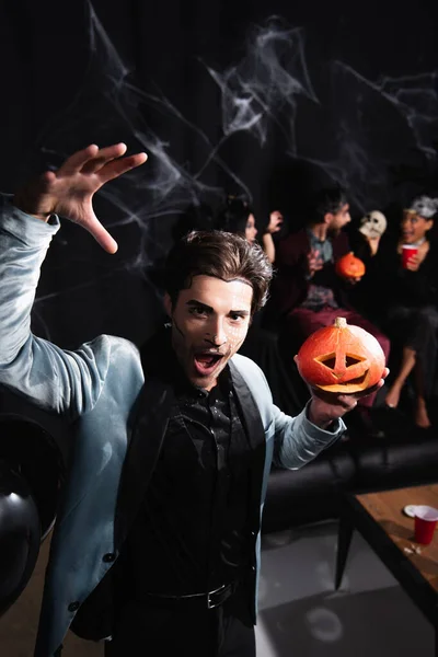 Uomo in Halloween trucco tenendo zucca intagliata, mostrando gesto spaventoso e ringhiando sul nero — Foto stock