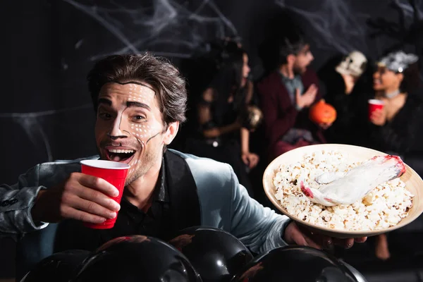 Happy man in halloween makeup drinking beer near balloons and blurred friends on black background — Stock Photo