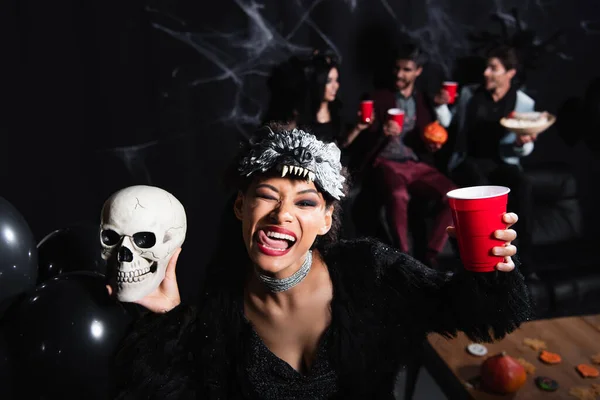 African american woman in wolf halloween mask sticking out tongue while holding spooky skull and plastic cup on black — Stock Photo