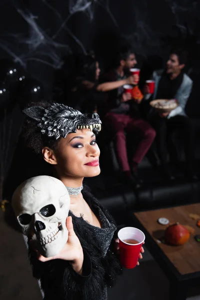 Femme afro-américaine souriante en loup-garou masque d'Halloween montrant crâne effrayant sur noir — Photo de stock