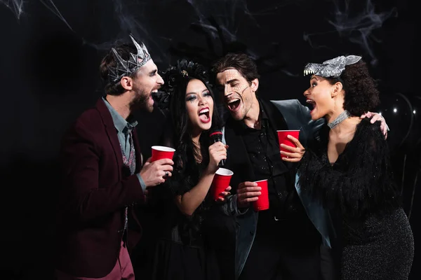 Amigos multiétnicos emocionados en disfraces de Halloween cantando karaoke en negro - foto de stock