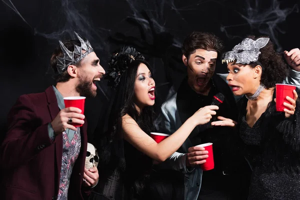 Alegres amigos multiétnicos con copas de plástico cantando karaoke durante la fiesta de halloween en negro - foto de stock