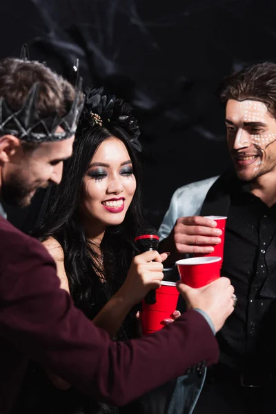 Улыбающаяся азиатка в костюме вампира на Хэллоуин поет караоке рядом с мужчинами на черном — стоковое фото