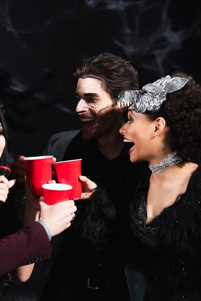 Excité afro-américaine femme cliquetis tasses en plastique avec des amis chantant karaoké sur fête d'Halloween sur noir — Photo de stock