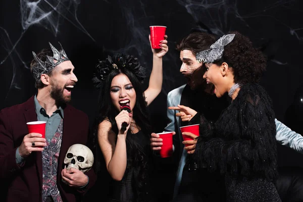 Mujer asiática con taza de plástico cantando karaoke durante la fiesta de Halloween con amigos multiétnicos en negro - foto de stock