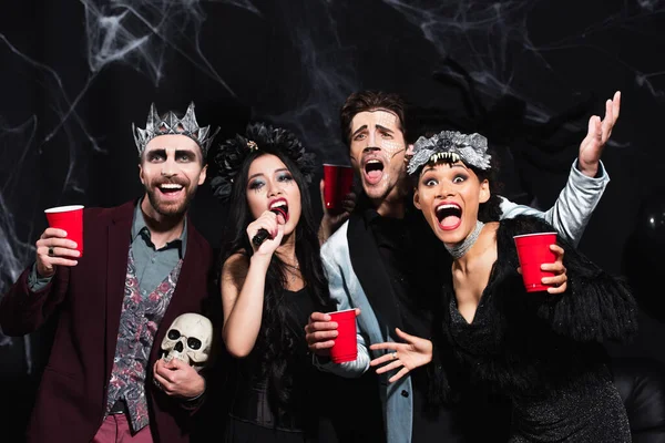 Cheerful multicultural friends in halloween makeup and costumes singing karaoke on black — Stock Photo