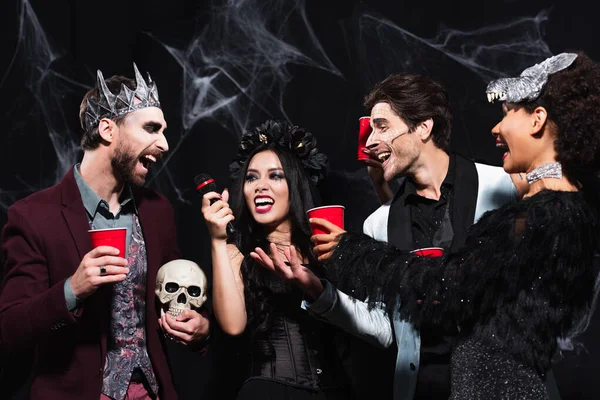 Glückliche asiatische Frau im Vampir-Halloween-Kostüm singt Karaoke in der Nähe multiethnischer Freunde auf schwarz — Stockfoto