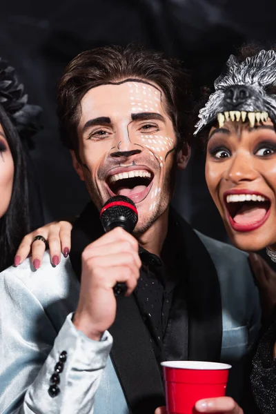 Man in halloween makeup singing karaoke near excited african american woman in wolf mask on black — Stock Photo