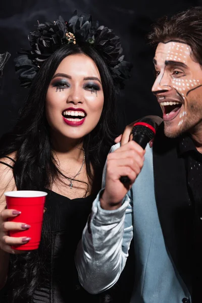 Feliz asiático mulher segurando plástico copo perto homem no halloween maquiagem cantando karaoke no preto — Fotografia de Stock