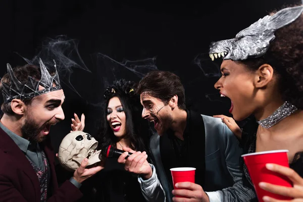 Hombre en halloween maquillaje celebración de micrófono cerca de calavera de miedo mientras canta karaoke con amigos multiétnicos en negro - foto de stock