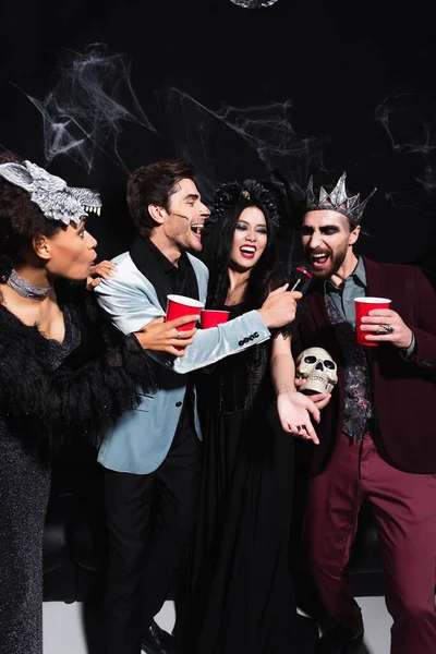 Hombre con cráneo y taza de plástico cantando karaoke cerca de amigos multiétnicos en trajes de Halloween en negro - foto de stock