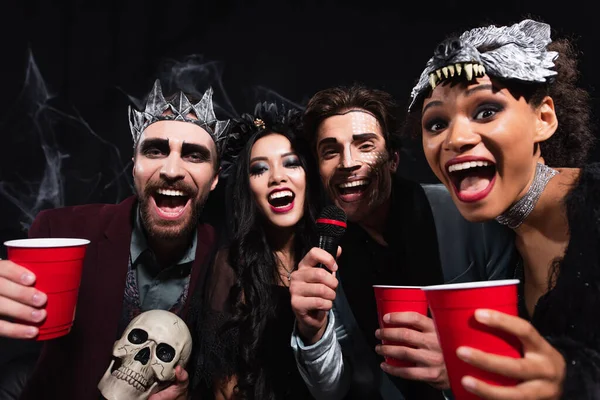 Взволнованные мультиэтнические друзья смотрят в камеру и поют караоке на вечеринке в честь Хэллоуина на черном — стоковое фото