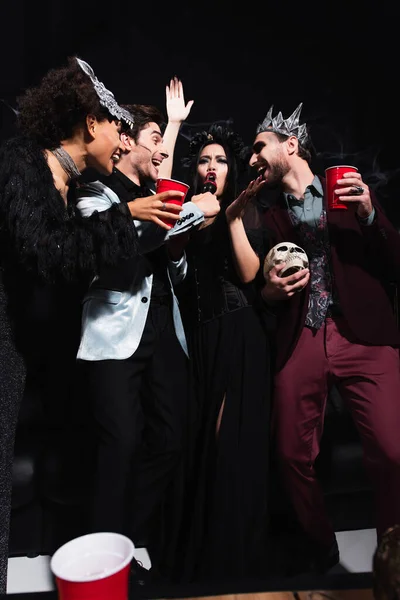 Збуджена азіатська жінка в костюмі вампіра Хеллоуїн співає караоке біля багатоетнічних друзів на чорному — стокове фото