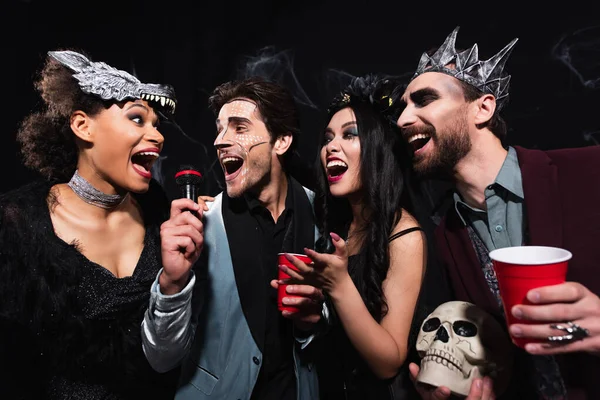 Joyful multiethnic friends in spooky costumes singing karaoke on halloween party on black — Stock Photo